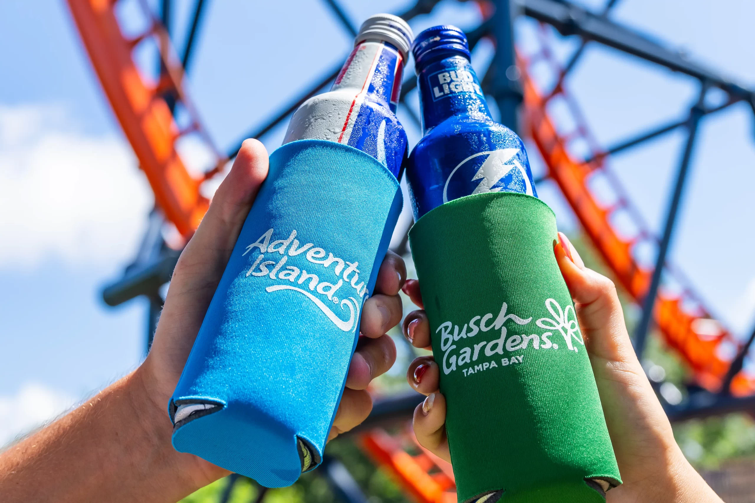 A blue Adventure Island can cooler and a green Busch Gardens Tampa Bay can cooler held up in front of a roller coaster