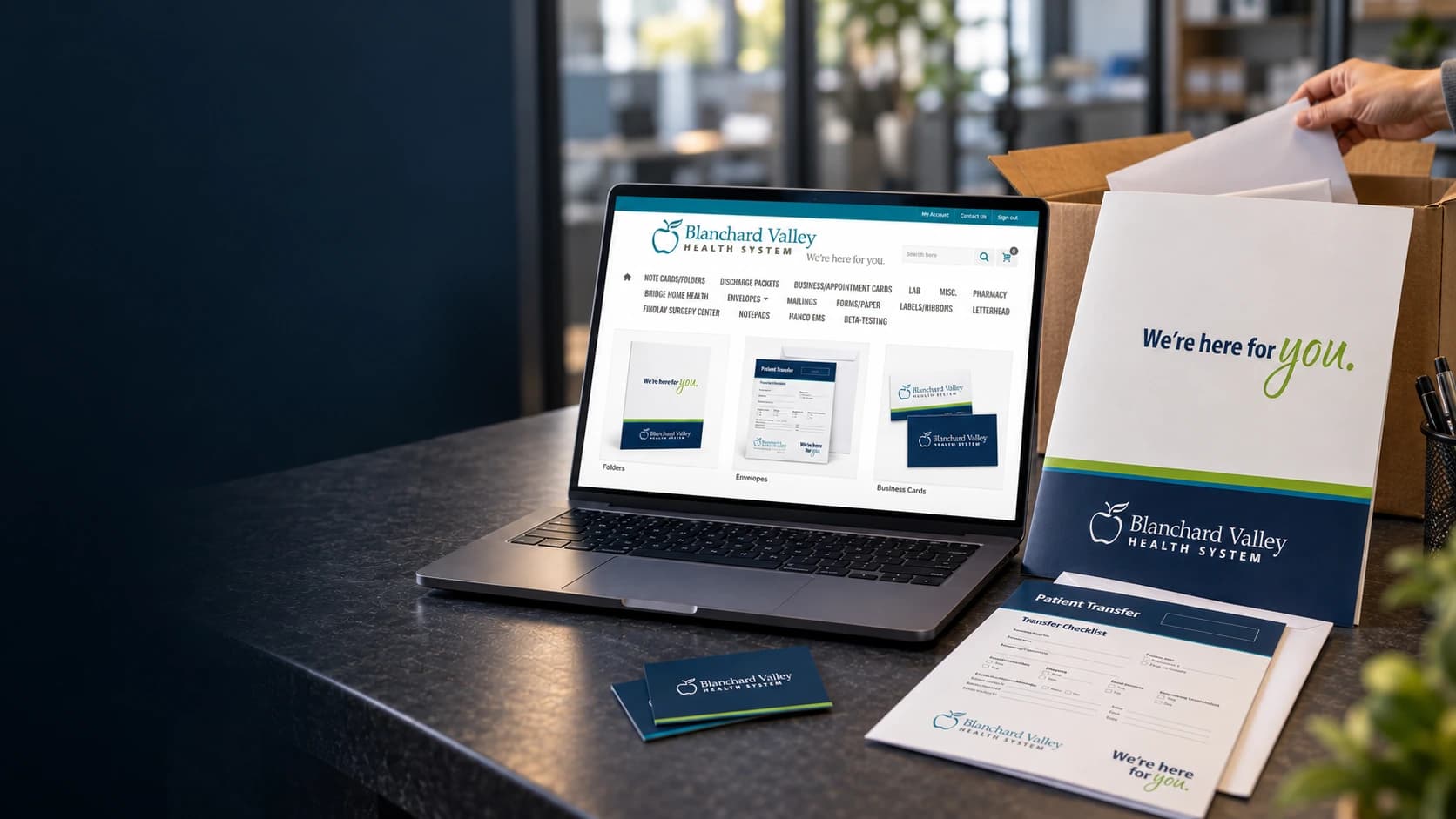Cinematic artwork of a laptop showing the Blanchard Valley Health System print store beside a presentation folder, a patient transfer envelope, and business cards