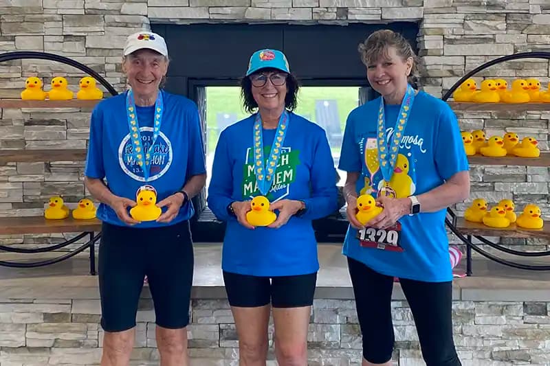 Three Mimosa 5K finishers in matching blue event shirts holding rubber ducks and wearing their medals