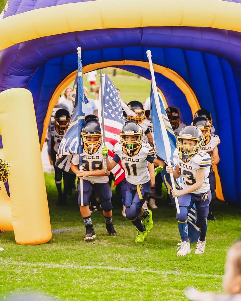 Young football players carrying flags as they run out of a giant blue and gold inflatable tunnel onto the field