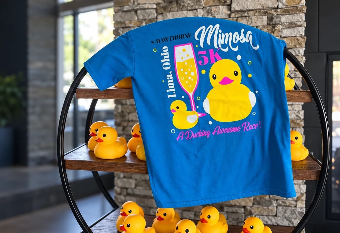 Cinematic artwork based on a customer photograph of a blue 19 Hawthorne Mimosa 5K shirt draped over a circular shelf with yellow rubber ducks