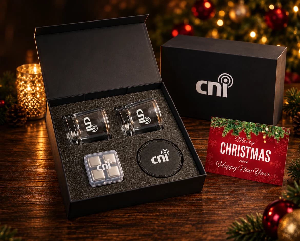 Cinematic artwork based on CNI's original product proof: the black whiskey gift set open on a wooden table, with two logo glasses, the ice cube case, a coaster, the logo gift box, and a red Christmas card