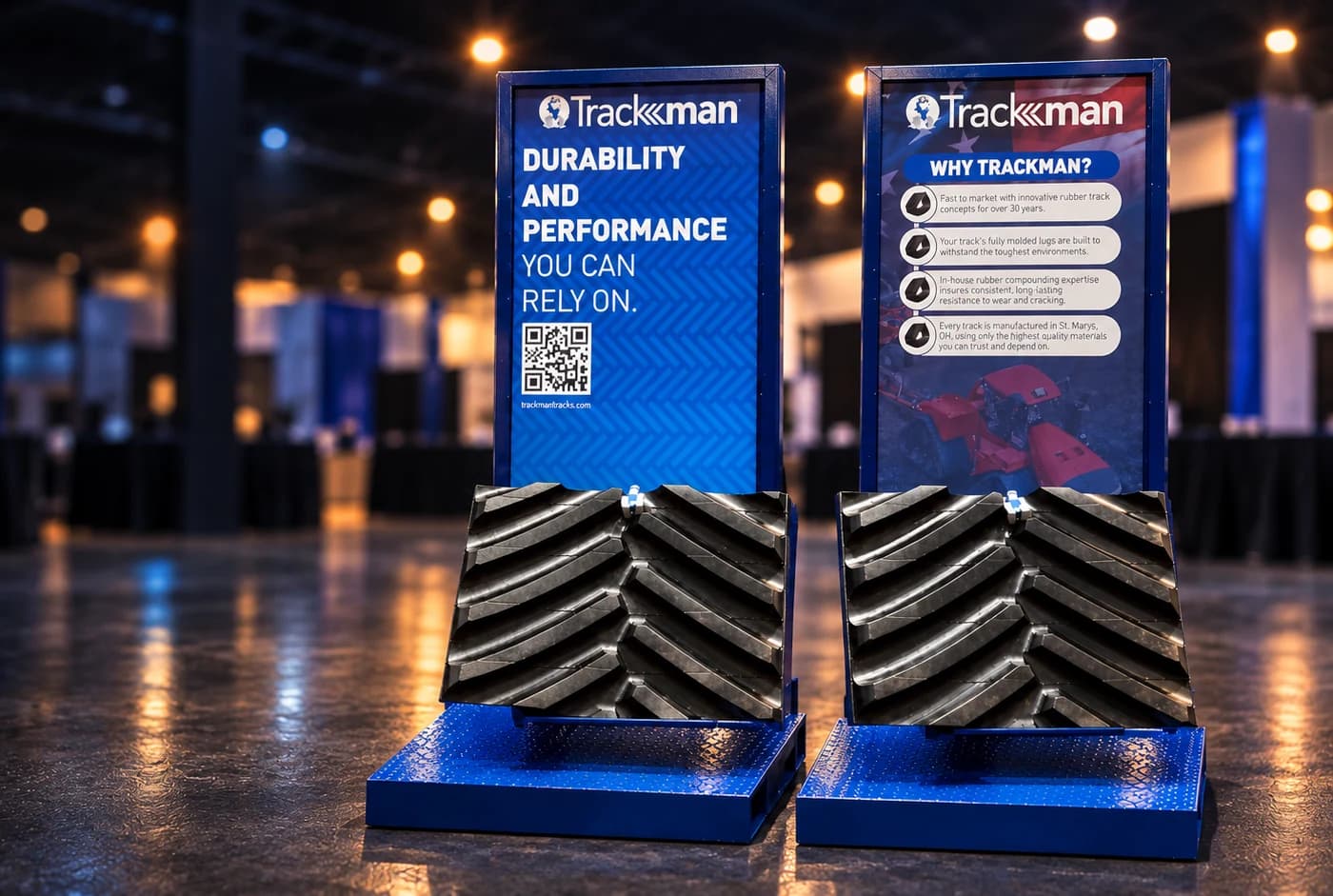 Cinematic artwork, based on photos of the actual displays, of two custom Trackman stands with blue frames and printed panels holding real sections of rubber track in an exhibition hall