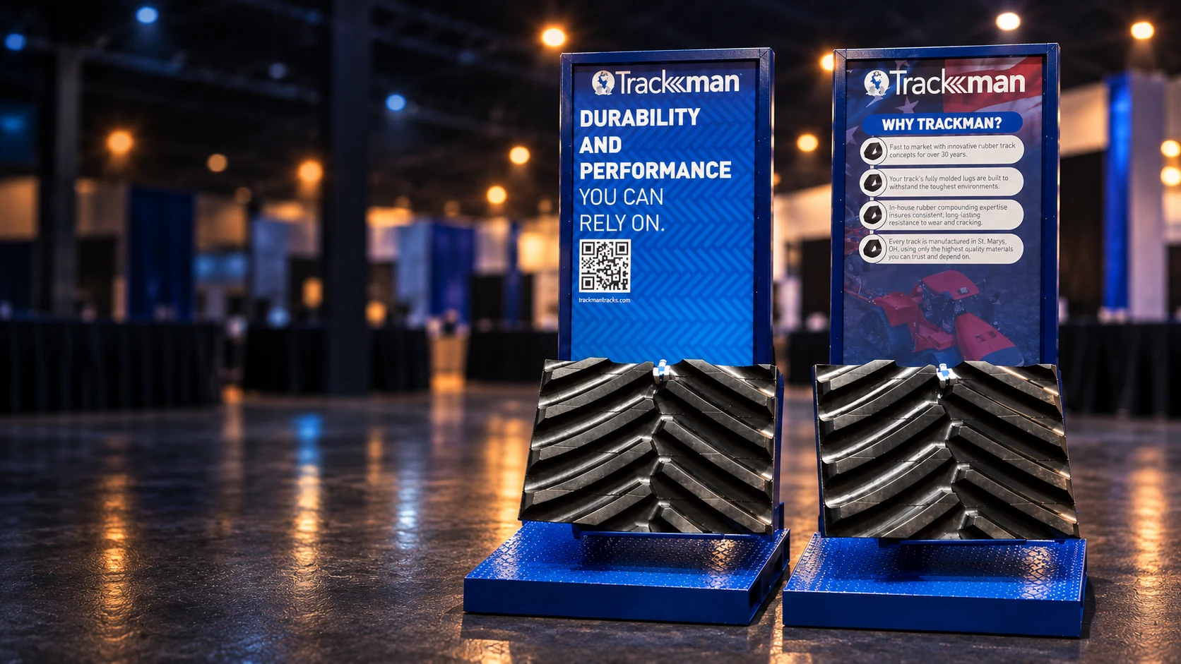 Cinematic artwork, based on photos of the actual displays, of two custom Trackman stands with blue frames and printed panels holding real sections of rubber track in an exhibition hall
